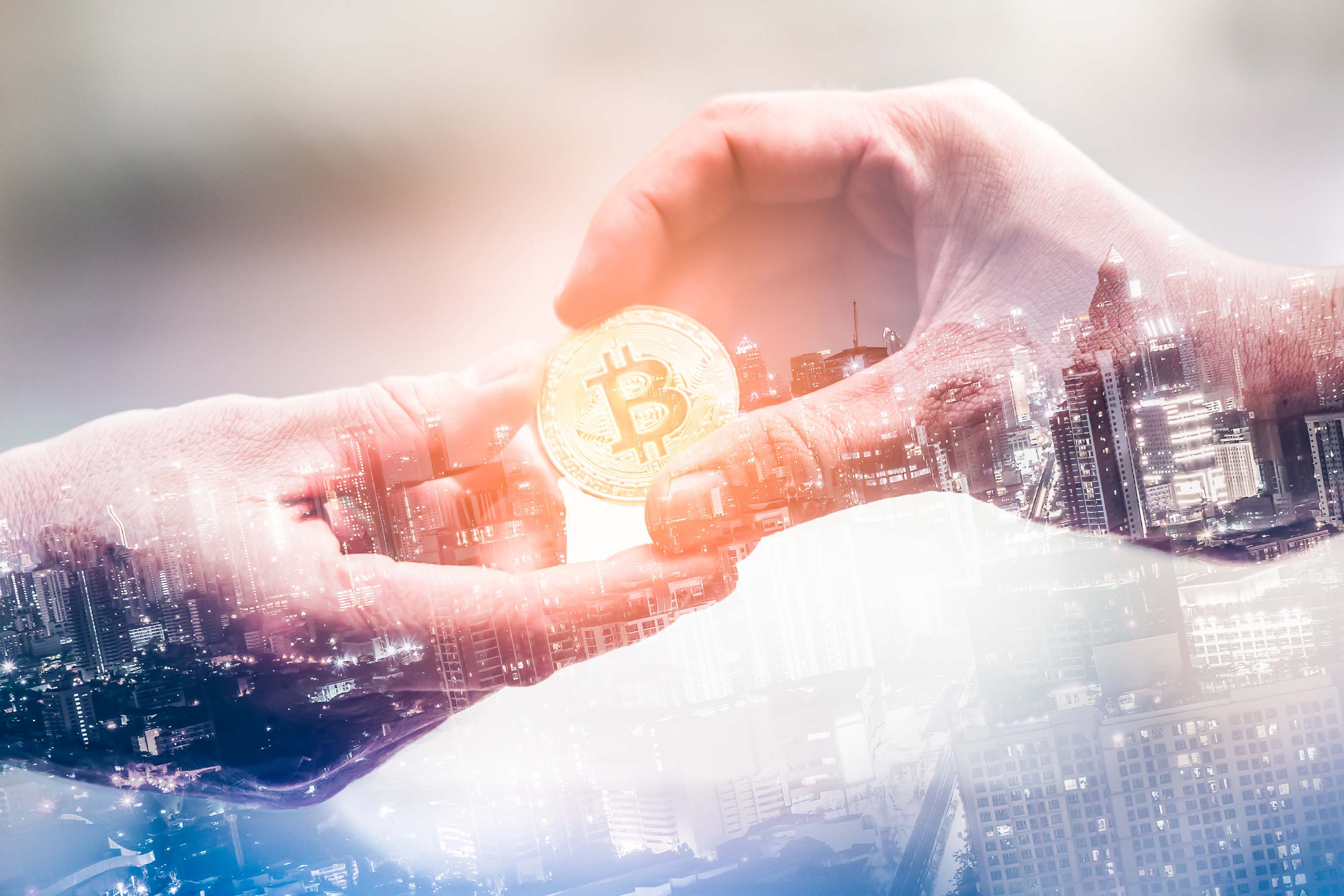 A composite image using double exposure technique: Two hands meet in the center, passing a gold Bitcoin coin from one to the other. This action is overlaid onto a background photograph of a brightly lit city skyline at night.